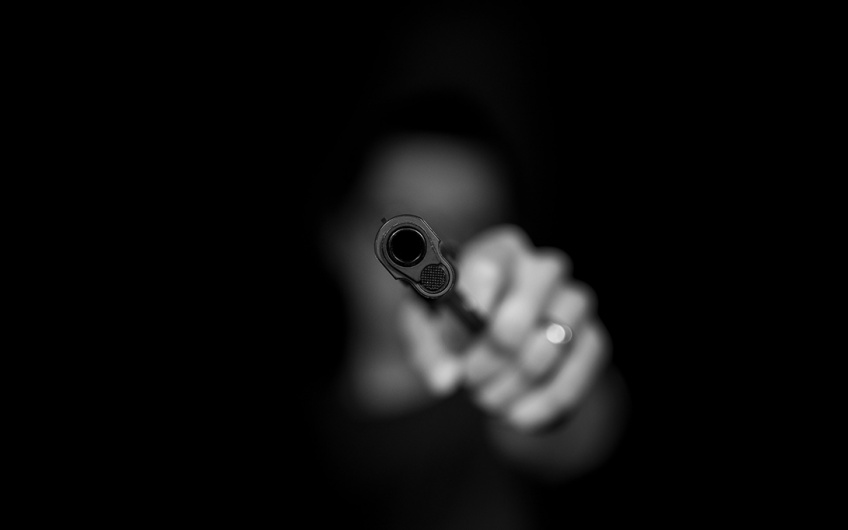 Black and white photo of a person pointing a firearm at the camera, the gun obscuring their face.
