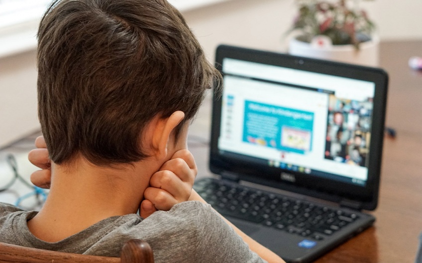 Photo of a young child facing away from the camera and looking into a laptop computer screen, their arms held against their chest with their hands folded up below their ears 