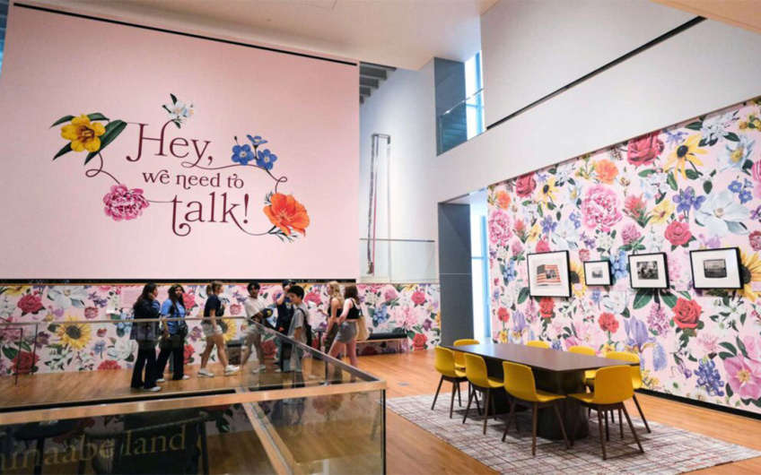 Students wander a room with framed artworks on floral wallpaper. The left wall reads in a decorative script, "Hey, we need to talk!"