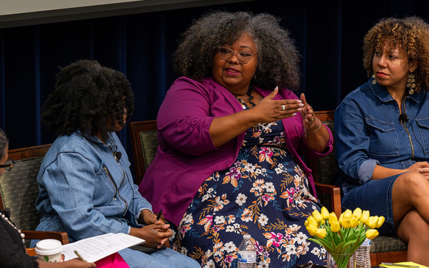 Four individuals participate in a panel discussion in a seminar room, with one speaker gesturing while talking. A bouquet of yellow tulips is on the table in front of them.