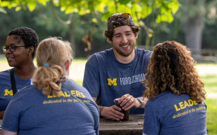 Group of four people wearing University of Michigan Ford School shirts, seated outdoors, engaged in a cheerful conversation.