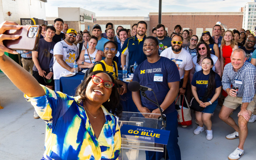 A group of fans at a University of Michigan event gather for a group selfie, with a person in the foreground holding out a phone. Everyone is wearing casual clothing, many sporting school colors and logos. A "Go Blue" banner is visible in the scene.