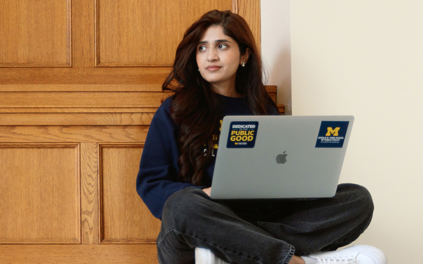 Person sitting with a laptop open on their lap, against a wooden wall. The laptop has a University of Michigan and an Apple logo sticker.