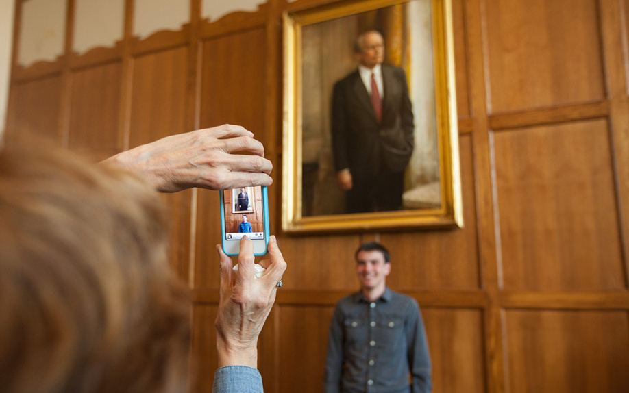 A person takes a photo with a smartphone of another person standing and smiling under a framed portrait of Gerald R. Ford in a wood-paneled room.