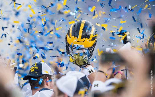 A Michigan football helmet held up above a crowd of players amid blue and maize confetti in the air