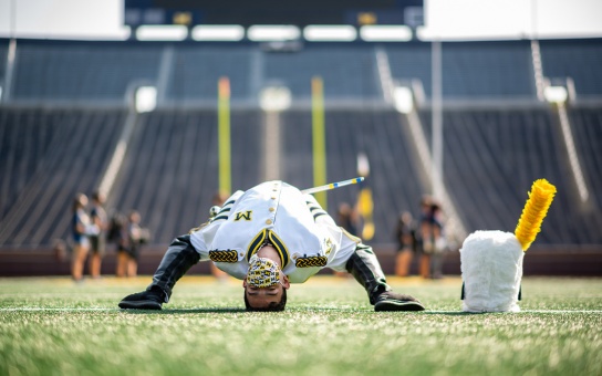 Photo of Walter Aguilar, in full drum major attire, bending backward to touch his head to the turf in Michigan Stadium
