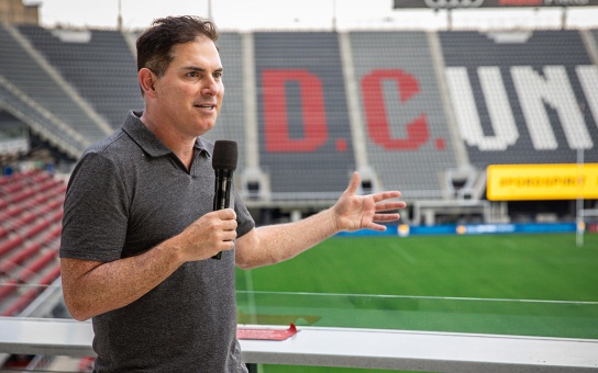 Photo of Jason Levien speaking into a microphone in Audi Stadium, with the stands in the background emblazoned with the DC United wordmark