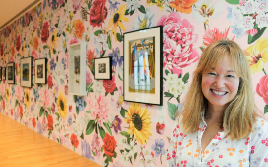 Phillipa Hughes (a white woman with shoulder length dirty blonde hair) stands smiling in the right third of the photo. She is shown from chest up with a floral shirt. Behind her is the wall of a hallway, with framed out-of-focus images on floral wallpaper.