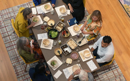 an overhead view of eight people sitting and conversing around a table laden with food and flyers titled with the name of the exhibit. 