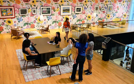 Students wander around a room with framed images on floral wallpaper. In the room there are various chair and sitting spaces, including a a table where some students sit and talk.