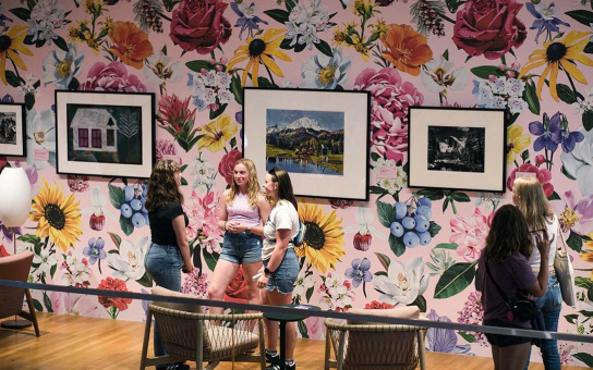 Three students stand and talk in front of a wall lined with framed artwork on top of floral wallpaper. Two others on the right of the image wander the exhibit.