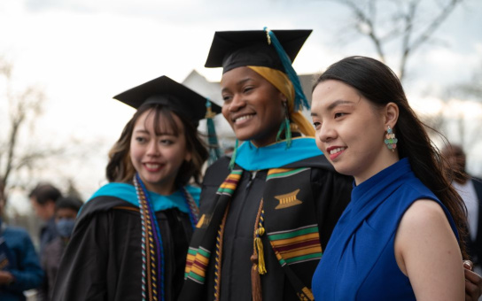 Three graduates in academic regalia, smiling joyfully during a graduation ceremony. The person in the center wears a black cap and gown adorned with a colorful stole, flanked by two individuals, one also in cap and gown and the other in a formal blue outfit.