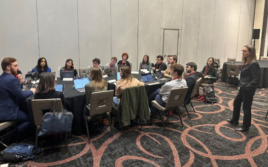 Group of attendees at a conference, seated around tables with laptops, paying attention to a speaker standing at the front of the room.