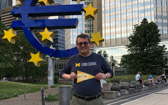 A person holding a sign that reads "M Ford School 50," standing in front of the large Euro sculpture outside the European Central Bank in Frankfurt.