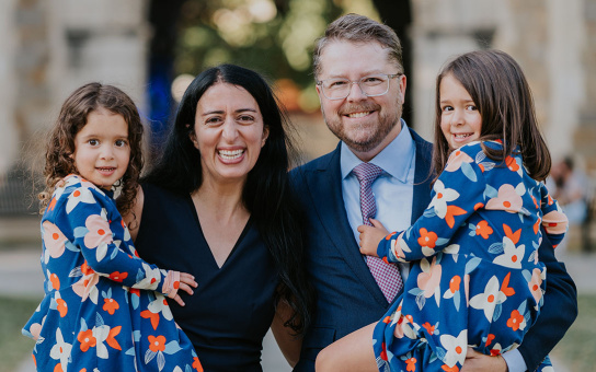 Two adults and two children smiling in a park, with the adults holding the children. The children are wearing matching floral dresses.