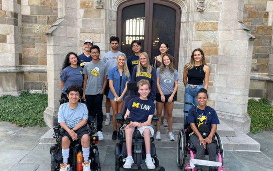 A group of students wearing University of Michigan attire poses in front of a historic building entrance. Some students are seated in wheelchairs.