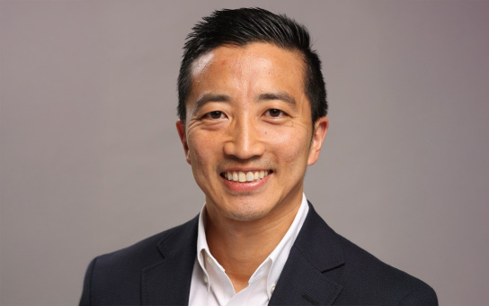 Portrait of a smiling individual in a business suit, against a neutral grey background.