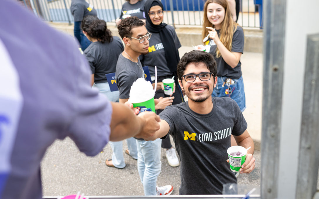 A smiling, PPIA fellow wearing a grey Ford School t-shirt reaches out to receive a cup of Kona Ice