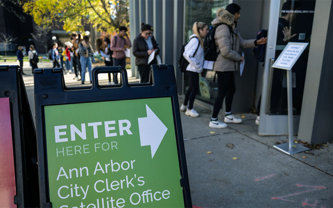 Students wait in line outside of UMMA on a sunny day. There is a green sign that reads "Enter here for Ann Arbor City Clerks Satellite Office."