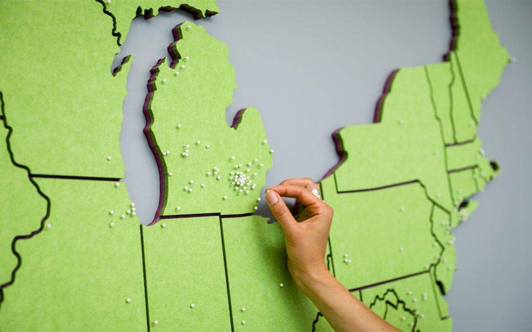 A hand places a pin in the Ann Arbor area on a green map of the US, zoomed in on Michigan.