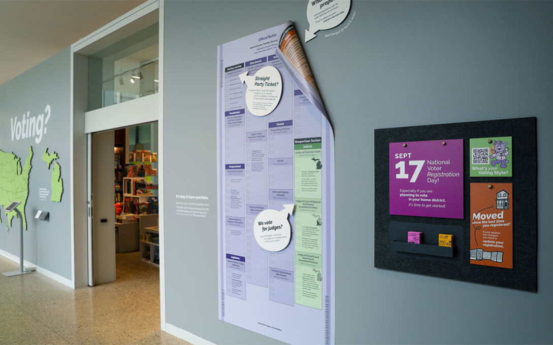 A hallway outside the gift shop in the University of Michigan Museum of Art. There are large wall displays of a US map, a ballot, and a spot to pick up information. Tables and chairs are on the opposite side of the hallway.