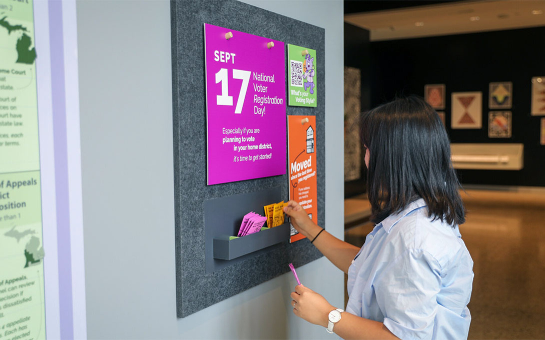 A student, in a button down shirt with shoulder-length dark hair, interacts with the brochures on the wall.