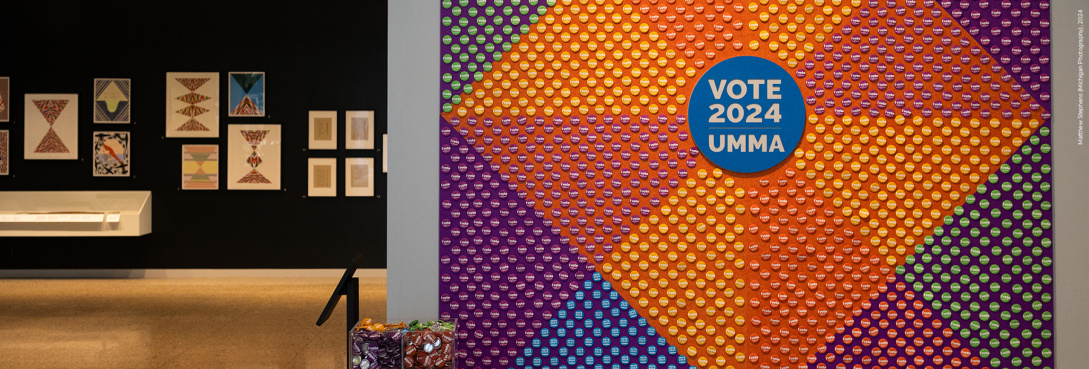 Thousands of small round buttons create a geometric pattern on a large, square, multi-colored mural, which hangs on a gray wall. At the center of the mural is a large blue circle that reads: “Vote 2024 UMMA.” A small white podium stands to the left of the mural on which a clear box sits, filled with buttons identical to those that compose the wall-mural.
