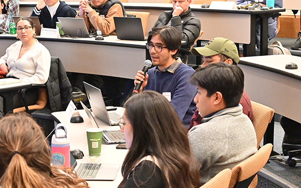 A student with black hair and a blue sweater speaks into a microphone from his desk while his peers listen attentively.