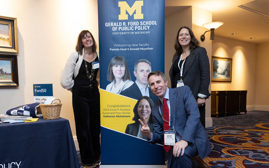 Four individuals pose at the Gerald R. Ford School of Public Policy event at the University of Michigan. They stand in front of a display featuring the university's logo and informational banners about the event.