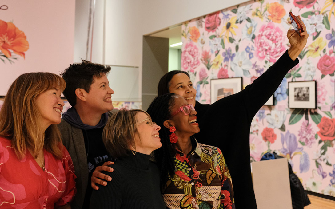 Group of five people taking a selfie in a room with floral wallpaper and framed photographs in the background.
