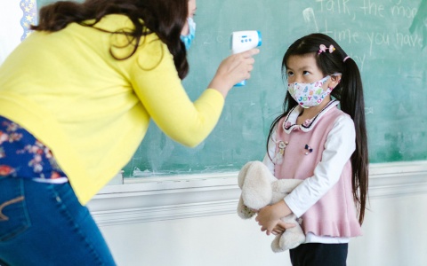 A teacher takes the temperature of a masked young student next to the class blackboard