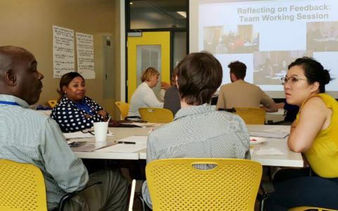 A group of adults sitting around a table in an office setting, having a conversation. Another table with a group can be seen behind them, as well as a PowerPoint slide that says "Reflecting on Feedback: Team Working Session."