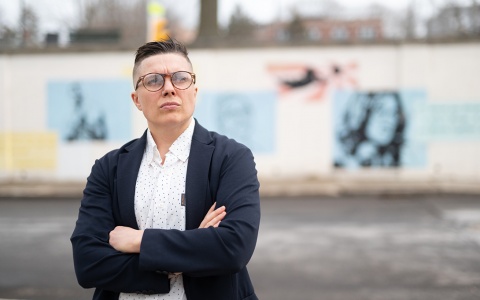 Kade Crockford, wearing glasses, a white collared shirt with dark sweater, arms are crossed, background is blurred wall