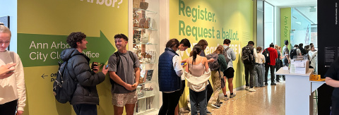Several college-aged students line up along the hallway of the University of Michigan’s Central Campus Voting Hub. The walls are painted light green with large white and dark green text that reads, “Voting in Ann Arbor?” and “Register, Request a ballot…” alongside some other text that is obscured by the queuing students. Two students waiting in line in the foreground, look at each other talking and smiling.