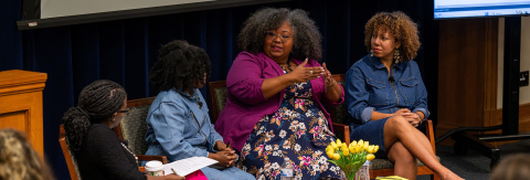 Four individuals participate in a panel discussion in a seminar room, with one speaker gesturing while talking. A bouquet of yellow tulips is on the table in front of them.