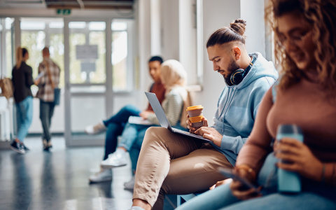 Photo of college students in a hallway, with focus on a student seated on a bench against the wall, holding a coffee and looking at their coffee.