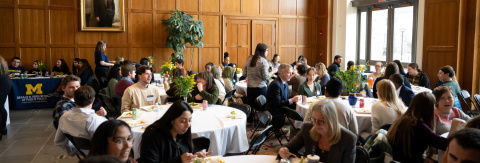 Group of people dining at a long table in a well-lit room with wooden panels and University of Michigan banners visible in the background.