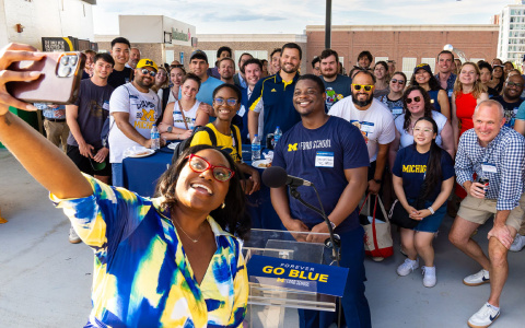 A group of fans at a University of Michigan event gather for a group selfie, with a person in the foreground holding out a phone. Everyone is wearing casual clothing, many sporting school colors and logos. A "Go Blue" banner is visible in the scene.