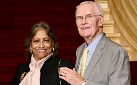 Two smiling individuals, one wearing a pink scarf and a badge, and the other in a light gray suit, standing together in front of a grand staircase.